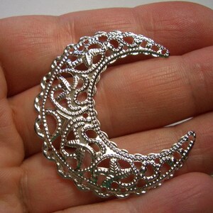 May include: A silver-toned crescent moon charm featuring an intricate, openwork filigree pattern with a scalloped edge. The metal piece has a reflective, polished finish and a curved shape that tapers to pointed ends, showcasing detailed scrollwork.