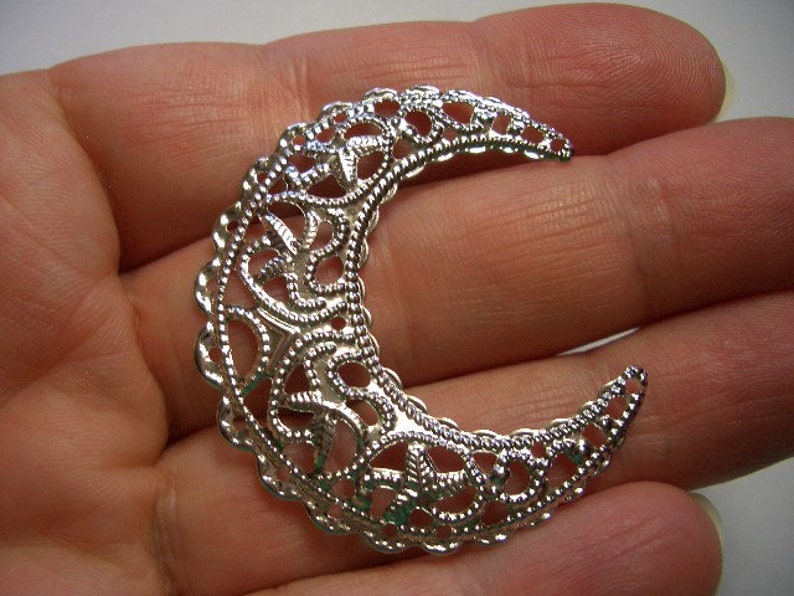 May include: A silver-toned crescent moon charm featuring an intricate, openwork filigree pattern with a scalloped edge. The metal piece has a reflective, polished finish and a curved shape that tapers to pointed ends, showcasing detailed scrollwork.