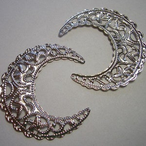 May include: Two silver-toned metal filigree crescent moon charms featuring an intricate, openwork lace-like pattern. The crescent shapes are curved with scalloped edges and a reflective, metallic finish, displaying a detailed, repeating decorative design.
