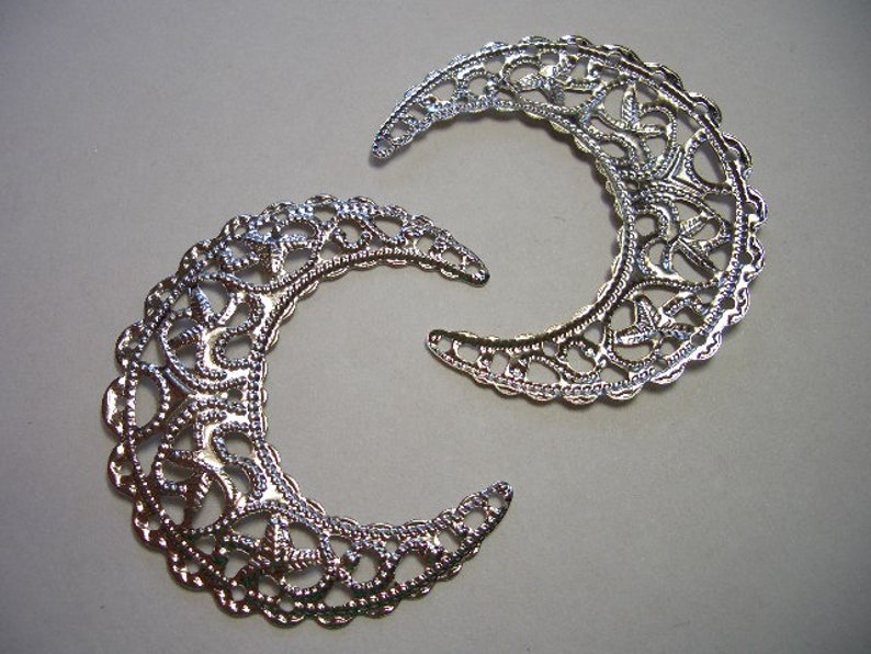 May include: Two silver-toned metal filigree crescent moon charms featuring an intricate, openwork lace-like pattern. The crescent shapes are curved with scalloped edges and a reflective, metallic finish, displaying a detailed, repeating decorative design.