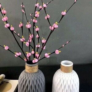 May include: Two decorative vases, one grey and one white, with textured surfaces and rope detailing around the necks. The grey vase holds branches with pink and yellow flower buds. The vases are on a dark surface.