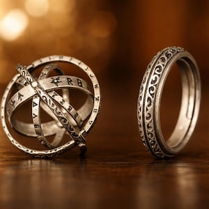 Peut inclure: Deux bagues en argent sont présentées sur une surface en bois. L'une est une sphère avec des bandes entrecroisées, l'autre une bague ornée d'un motif. L'arrière-plan est flou avec des tons chauds.