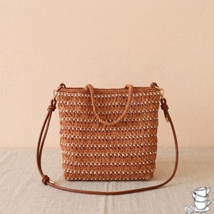 Op de afbeelding: Een bruin en wit gehaakte tas met een textuur van horizontale strepen. De tas heeft twee korte handvatten en een lange, dunne, verstelbare bruine schouderriem die aan beide zijden is bevestigd met goudkleurige metalen hardware.