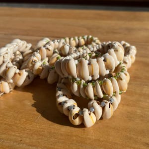 May include: Several bracelets made of small, cream-colored seashells. The shells are strung together with silver-colored beads and small green beads. The bracelets are arranged on a wooden surface.
