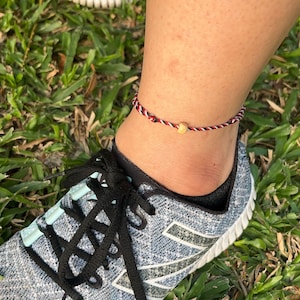 May include: Ankle bracelet with red, white, and black braided cord and a small wooden bead. The bracelet is worn on an ankle above a gray and blue athletic shoe with black laces, set against a background of green grass.