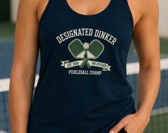 Könnte beinhalten: Marineblaues Tanktop mit dem Text "DESIGNATED DINKER" über einer Grafik von Pickleball-Schlägern und einem Ball. Darunter steht "EST. 2026 THE KITCHEN PICKLEBALL CHAMP" auf einem Banner. Das Tanktop ist ärmellos.