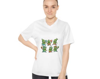 Könnte beinhalten: Weißes V-Ausschnitt-T-Shirt mit einer Cartoon-Grafik von grünen Gurken-Charakteren, die Pickleball spielen. Die Gurken tragen verschiedene Outfits und halten Schläger. Das T-Shirt wird von einer Person getragen.