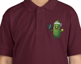 Könnte beinhalten: Burgunderfarbenes Poloshirt mit einem Cartoon-Gurken-Charakter, der Pickleball spielt. Die Gurke ist grün mit einem weißen Stirnband, hält einen Schläger und einen gelben Ball. Das Shirt hat einen Kragen und Knopfverschluss.