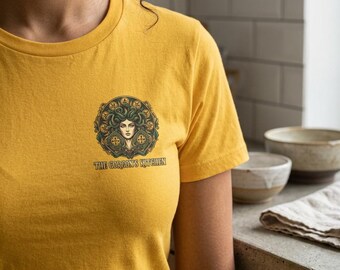 Könnte beinhalten: Ein senfgelbes T-Shirt mit einer kreisförmigen Grafik auf der Brust. Das Design zeigt eine stilisierte weibliche Figur mit schlangenartigem Haar, umgeben von verzierten grünen und goldenen Mustern. Unter der Grafik steht der Text THE GORGON'S KITCHEN.