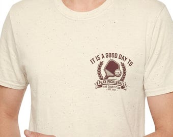 Könnte beinhalten: Cremefarbenes T-Shirt mit dem Text "IT IS A GOOD DAY TO PLAY PICKLEBALL" über einem Pickleball-Schläger- und Ball-Design. Darunter steht "THE COURT CLUB - EST. 2021".