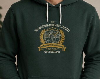 Könnte beinhalten: Dunkelgrüner Kapuzenpullover mit weißem Kordelzug. Auf der Vorderseite befindet sich ein gold-weißes Emblem mit dem Text "The Kitchen-Avoidance Society" und "Embracing the Power Game". Das Emblem enthält eine Pickleball-Illustration und die Worte "Pure Pickleball".