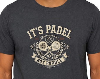 Könnte beinhalten: Dunkelgraues T-Shirt mit einem verblassten Aufdruck "IT'S PADEL NOT PADDLE". Das Design zeigt zwei Padel-Schläger, einen Ball und den Text "PCC 2025". Das Shirt hat einen Rundhalsausschnitt.