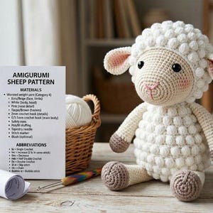 May include: A handmade white and beige amigurumi sheep sits next to a basket of yarn and a crochet hook. The sheep has a textured body, pink cheeks, and black safety eyes. A pattern sheet lists materials and abbreviations.