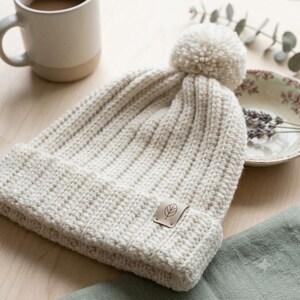 May include: A cream-colored knit beanie featuring a ribbed texture, a large pom-pom on top, and a folded cuff. A small, square, light-colored leather tag is attached to the cuff, displaying a simple line-art drawing of a single leaf.