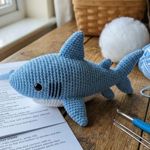 Könnte beinhalten: Ein gehäkeltes blaues Hai-Spielzeug mit weißem Bauch und schwarzen Augendetails. Der Hai hat Flossen und einen Schwanz und sitzt auf einer Holzoberfläche. Ein Häkelhaken und Garn befinden sich im Hintergrund.