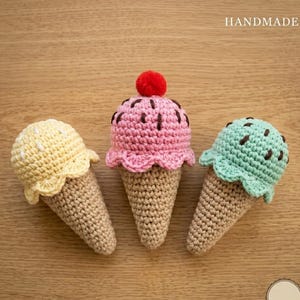 Op de afbeelding: Drie handgemaakte gehaakte ijshoorntjes in pastelkleuren. Eén is geel, één is roze met een rode pompon en één is mintgroen. De hoorntjes zijn beige. Het woord "HANDMADE" staat rechtsboven.