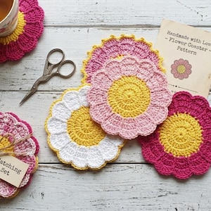Puede incluir: Posavasos de flores de ganchillo hechos a mano en tonos rosa, amarillo y blanco. Un posavasos está debajo de una taza de té, y otro juego está atado con hilo y una etiqueta que dice "Matching Set". También se ven unas tijeras y un patrón.