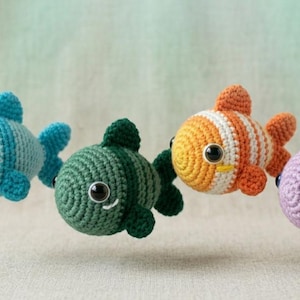 May include: Four small, round, crocheted fish toys in light blue, dark green, orange and white striped, and light purple. Each fish features a textured yarn surface, two dark plastic safety eyes, and small crocheted fins and tails.