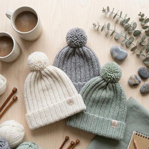 May include: Three knit beanies in cream, grey, and sage green, each featuring a ribbed texture, a matching pom-pom on top, and a small rectangular tag with a leaf icon on the folded cuff. The hats are arranged together on a light surface.