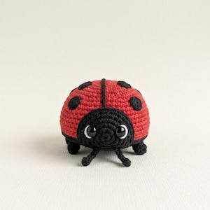 May include: A handmade crochet ladybug toy. The ladybug has a red body with black spots and a black head. It has small black legs and antennae, and large white-rimmed eyes. The toy is made of yarn and is sitting on a white surface.