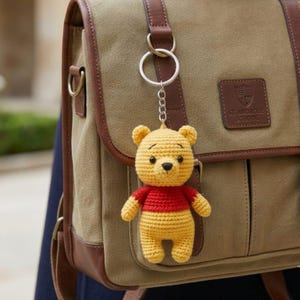 May include: A crochet keychain featuring a yellow bear with a red shirt attached to a silver metal ring. The bear has black eyes and a black nose. The keychain is clipped to a tan canvas backpack with brown leather trim and a patch that reads ST. ANDROSE SCHOOL.
