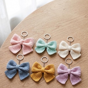May include: Six crochet bow keychains in various colours including light pink, mint green, cream, dusty blue, mustard yellow, and lavender. Each bow is attached to a silver-toned metal split ring by a short chain, featuring a textured yarn finish.