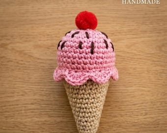 May include: A handmade crochet ice cream cone. The ice cream is pink with brown sprinkles and a red pom-pom cherry on top. The cone is beige. The word "HANDMADE" is in the top right corner.