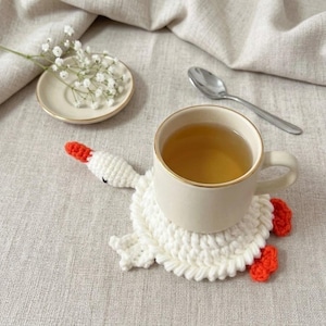 Peut inclure: Une tasse en céramique blanche remplie de thé repose sur un dessous de verre en crochet blanc fait main en forme d'oie avec un bec et des pattes orange. À côté se trouve une petite soucoupe blanche avec du gypsophile et une cuillère en métal.