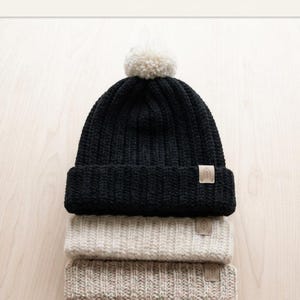 May include: A stack of three ribbed knit beanies. The top beanie is black with a white pom-pom and a small tan rectangular tag featuring a leaf icon. The middle beanie is cream, and the bottom beanie is a light tan, both with matching tags.