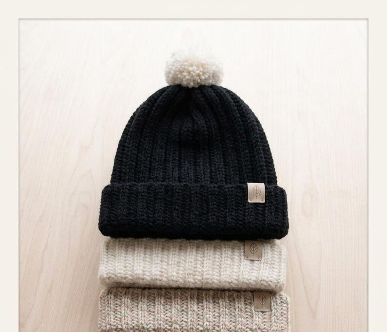 May include: A stack of three ribbed knit beanies. The top beanie is black with a white pom-pom and a small tan rectangular tag featuring a leaf icon. The middle beanie is cream, and the bottom beanie is a light tan, both with matching tags.
