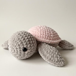 Puede incluir: Una tortuga de ganchillo hecha a mano con hilo suave y afelpado. La tortuga presenta una cabeza y extremidades grises, un caparazón rosa y ojos de seguridad negros. El peluche está diseñado con una superficie texturizada.