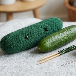 Può includere: Un cetriolo verde lavorato a maglia con occhi a bottone neri accanto a un cetriolo vero. Sono presenti anche ferri da maglia, filo verde e un ago, a suggerire un tema artigianale. Gli oggetti poggiano su una superficie grigia.