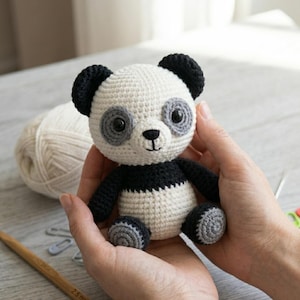 May include: A handmade crochet panda bear with black and white yarn. The panda has black ears, eyes, and limbs, with a white body and gray accents. The panda is being held in someone's hands.