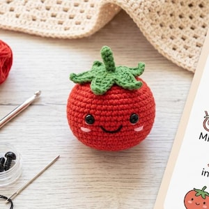May include: A handmade, red crocheted tomato with a smiling face and green leafy top. The amigurumi tomato is next to crochet supplies, including yarn, a crochet hook, and a needle. A guide with crochet instructions is also visible.