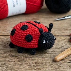 May include: A handmade, red and black crocheted ladybug sits on a wooden surface. The ladybug has black spots and a black head with antennae. Red yarn, black yarn, scissors, and a crochet hook are in the background.