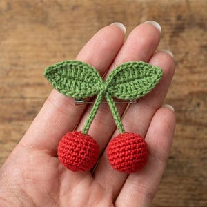 Peut inclure: Broche faite à la main en forme de deux cerises rouges avec des feuilles vertes. La broche est fixée avec une épingle argentée au dos. Les cerises sont rondes et texturées, et les feuilles sont détaillées avec des coutures visibles.