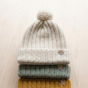 May include: A stack of three ribbed knit beanies, each featuring a small rectangular tag with a leaf icon. The top beanie is cream-colored with a matching pom-pom, the middle is sage green, and the bottom is mustard yellow. All have a folded cuff.