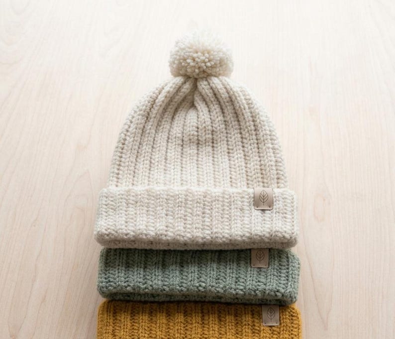 May include: A stack of three ribbed knit beanies, each featuring a small rectangular tag with a leaf icon. The top beanie is cream-colored with a matching pom-pom, the middle is sage green, and the bottom is mustard yellow. All have a folded cuff.