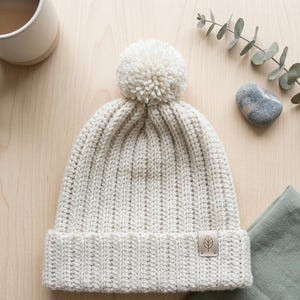 May include: A cream-colored knit beanie featuring a ribbed texture, a large matching pom-pom on the crown, and a folded brim. A small, rectangular, light brown leather-like tag with a simple leaf icon is attached to the lower right side of the brim.