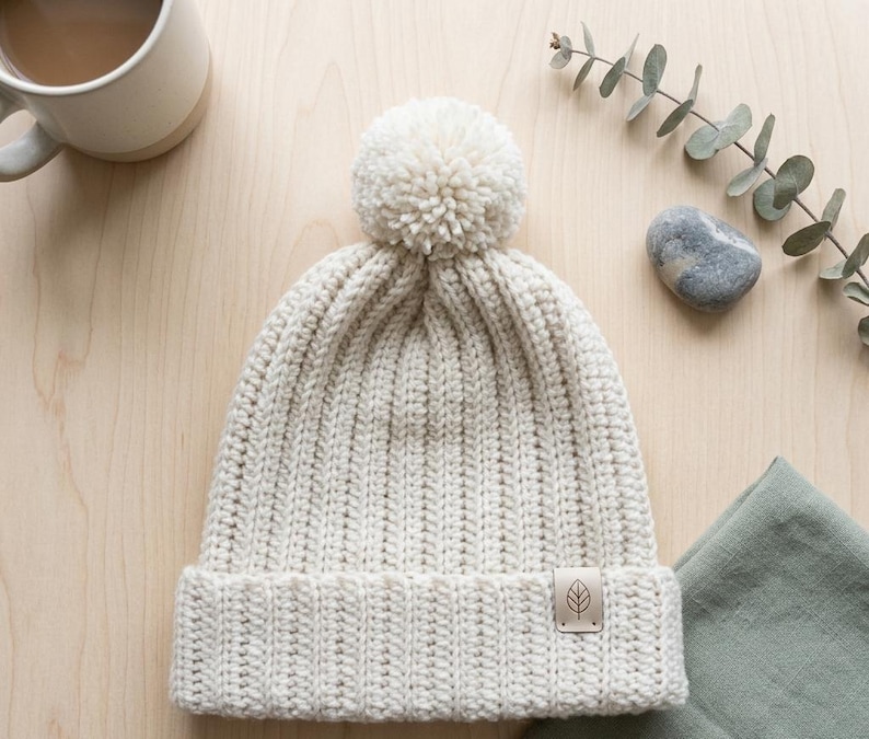 May include: A cream-colored knit beanie featuring a ribbed texture, a large matching pom-pom on the crown, and a folded brim. A small, rectangular, light brown leather-like tag with a simple leaf icon is attached to the lower right side of the brim.