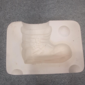 May include: A light-colored, rectangular mold featuring a detailed Santa Claus boot design. The boot has a textured appearance with visible stitching and decorative elements. The mold includes a rounded heel and a circular indentation.