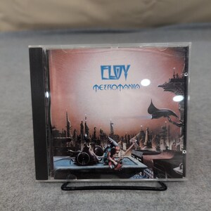 Puede incluir: Una caja de CD con el álbum "Metropomania" de Eloy. La portada muestra una ciudad futurista con una persona tocando la guitarra. El título del álbum y el nombre de la banda están en azul sobre un fondo rojizo-anaranjado.