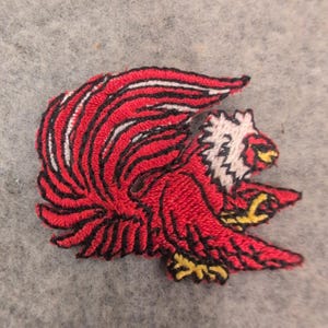 May include: An embroidered patch featuring a stylized eagle in profile. The bird has a bright red body and wings with black outlines, a white head with black detailing, and yellow talons. The design is rendered in dense thread stitching.