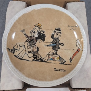 May include: A round white ceramic decorative plate featuring a tan center with a Norman Rockwell illustration titled Promenade à Paris. The artwork depicts three figures walking, with text reading Vote for Al, Egbert, and the artist's signature.