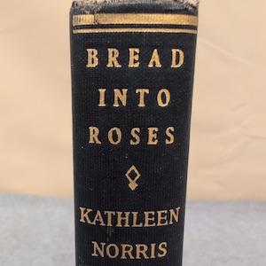 Puede incluir: Primer plano del lomo de un libro antiguo. El título "BREAD INTO ROSES" está en letras doradas, con el nombre del autor, "KATHLEEN NORRIS", debajo. El lomo es negro con un borde decorativo dorado en la parte superior.