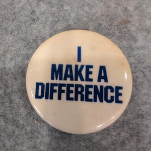 May include: A round, off-white pinback button featuring the text I MAKE A DIFFERENCE printed in bold, dark blue capital letters. The design is centered on the circular surface, with the word I on the top line and the remaining words below.