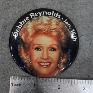 Op de afbeelding: Een ronde, zwarte knoop met een portret van Debbie Reynolds en de tekst "Debbie Reynolds • Jan. 16th". De afbeelding toont Reynolds lachend, met blond haar en oorbellen. De knoop heeft een diameter van ongeveer 5 cm.