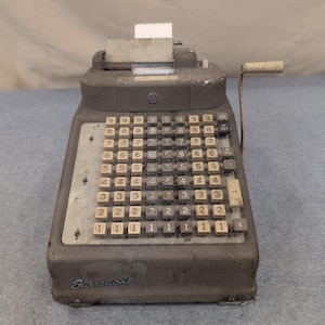 Op de afbeelding: Een antieke Burroughs-telmachine met een grijze metalen behuizing en talrijke genummerde toetsen. De machine heeft een slinger aan de zijkant en een papieren rol aan de bovenkant. Het woord "Burroughs" is zichtbaar.
