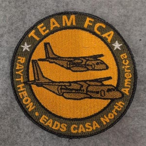 Puede incluir: Parche bordado circular con un centro dorado y dos aviones marrones. El anillo exterior es verde oliva y presenta el texto "TEAM FCA", "RAYTHEON", "EADS CASA North America" y dos estrellas blancas.