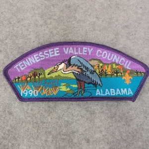 Pode incluir: Patch bordado com borda roxa e o texto "Tennessee Valley Council" e "Alabama". O patch apresenta uma garça azul em pé na água, com uma paisagem de árvores e o ano de 1990.